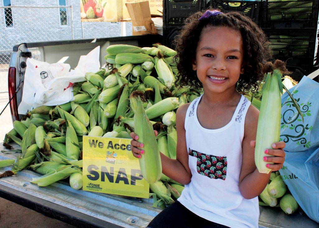 girl holding corn at the farmers market in front of sign that reads "we accept SNAP"