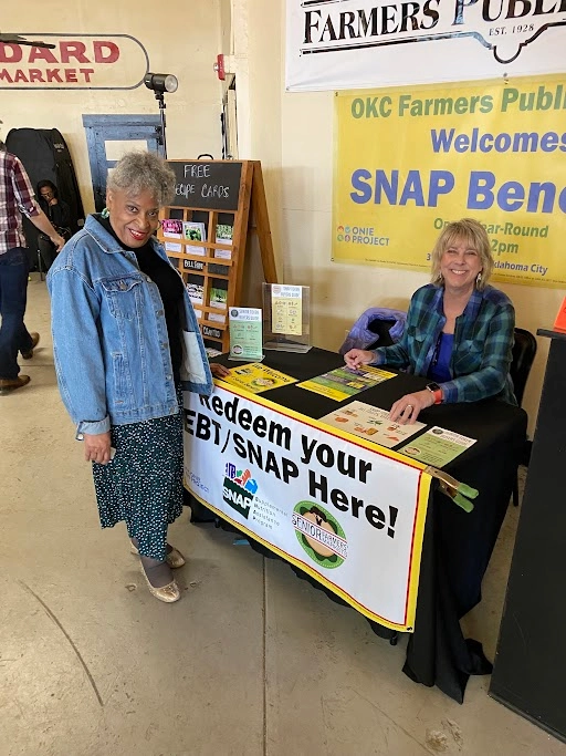 Two women standing in front of an EBT booth at a local farmers market