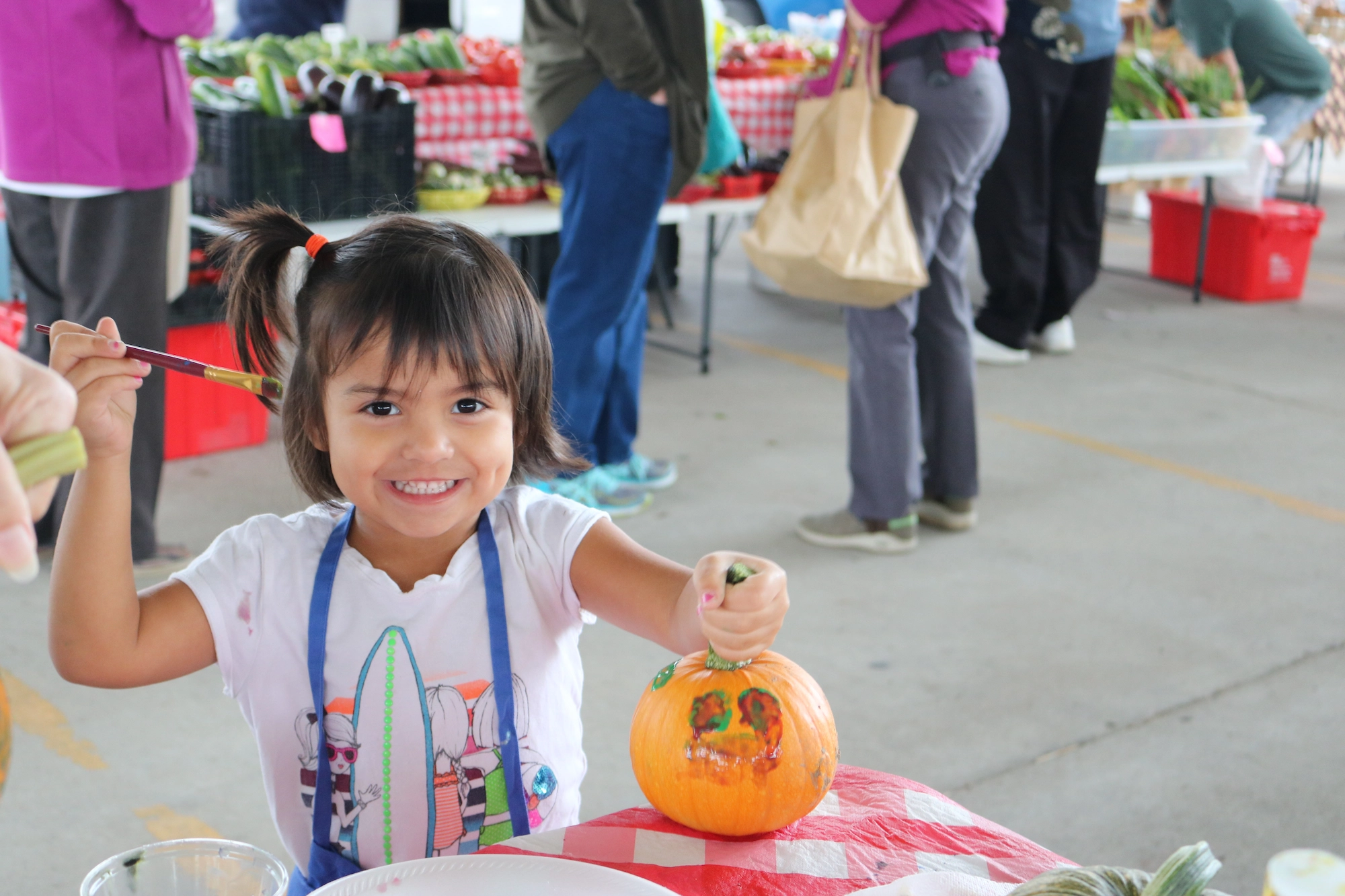 A grinning child presents her painted pumpkin for the camera at a farmers market. Shoppers can be seen in the background.