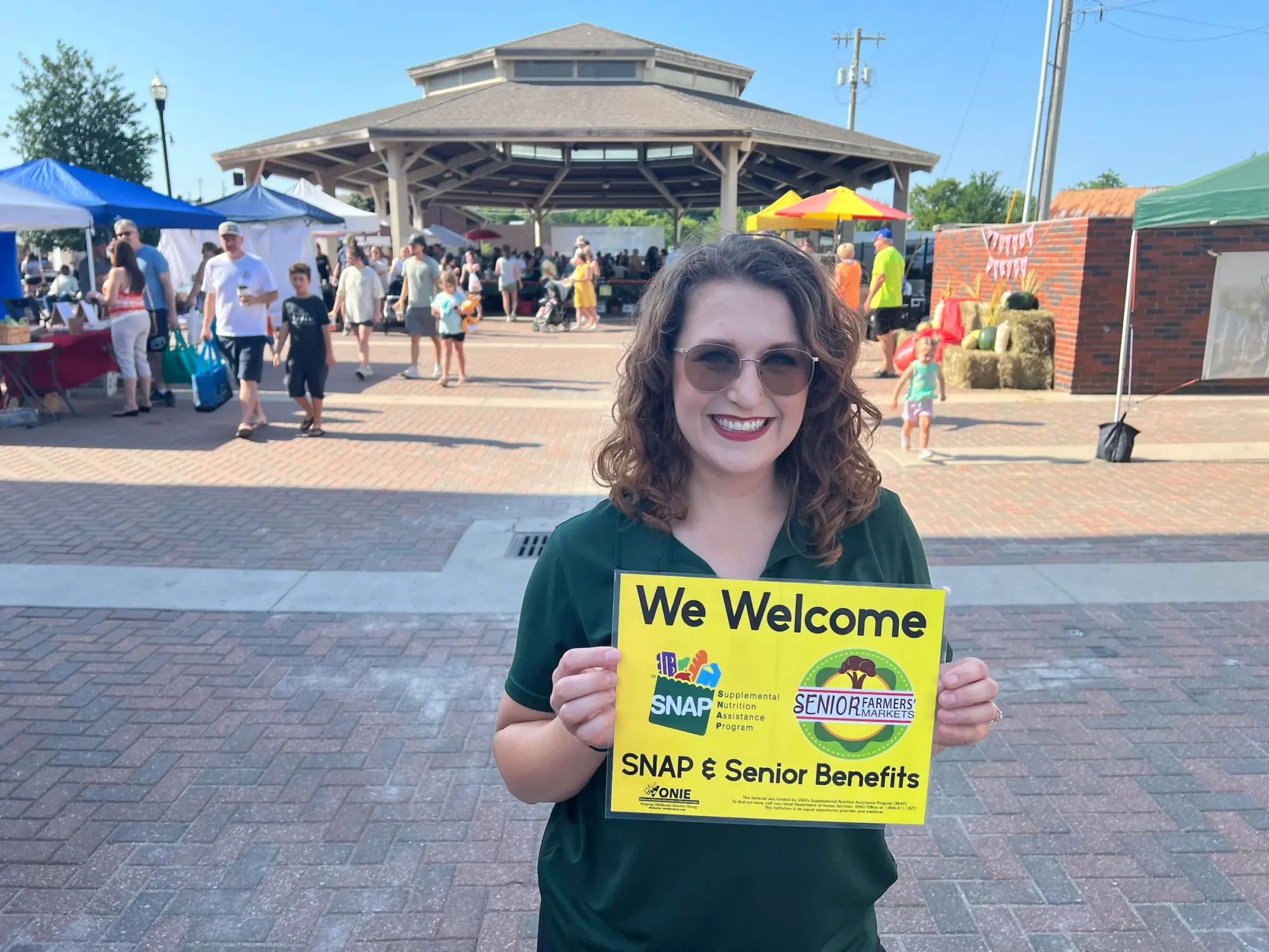 The Broken Arrow, OK Farmers Market manager holds up a yellow sign that says "We Welcome SNAP and Senior Benefits." The market, with it's tents and vendors, appears in the background.