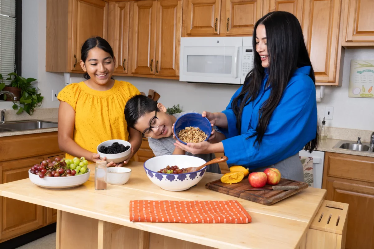 A woman is accompanied by two kids in the kitchen. They are smiling and prepping produce together.
